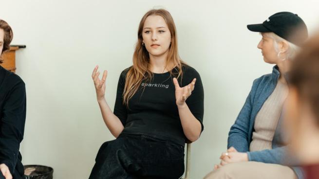 Kvinna med svarta kläder och långt hår gestikulerar för en grupp på tre andra