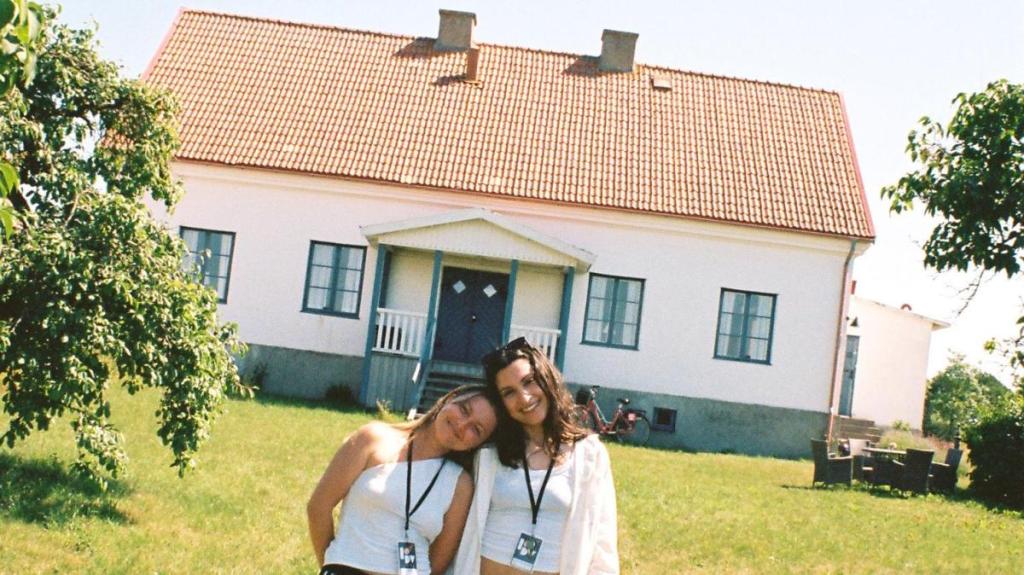 Två unga kvinnor i sommargrönskan framför mindre hus.