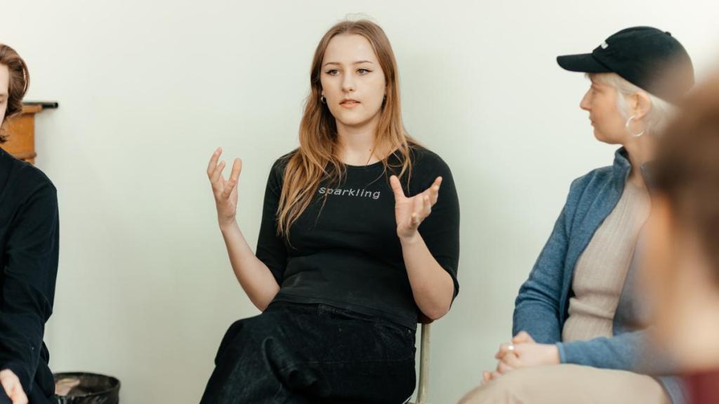 Kvinna med svarta kläder och långt hår gestikulerar för en grupp på tre andra