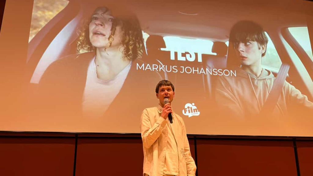 Man med vit skjorta framför en filmduk där bild på två personer i en bil projiceras.